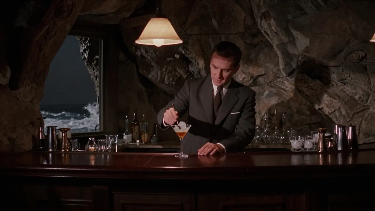 Intimate scene inside The Rock Bar, bartender stirring a martini in a stone-clad cave-like lounge with ocean sounds.